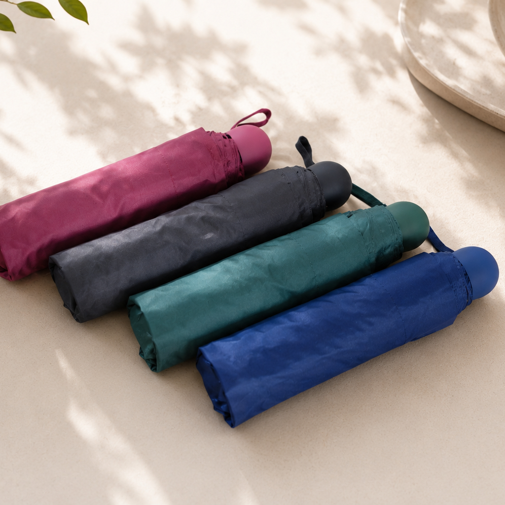 Four folded umbrellas in purple, blue, green, and black on a light surface with a plant and cup in the background.