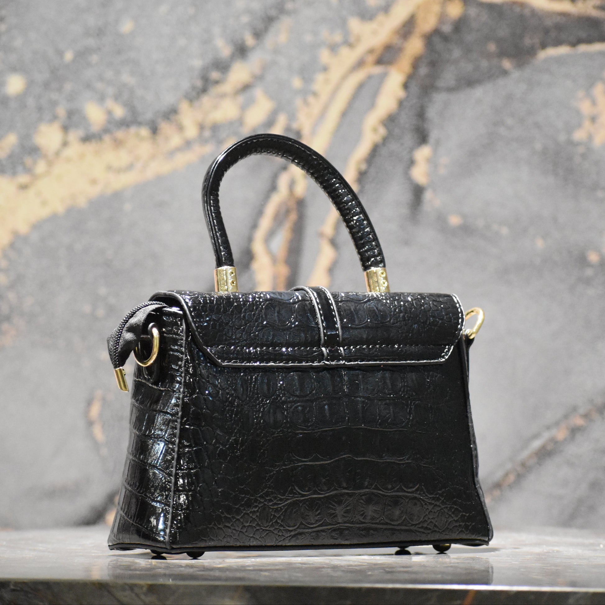 A structured black crocodile-embossed leather handbag with a top handle, gold-tone hardware, and small feet, displayed on a marble surface against a textured gray and gold marble background.